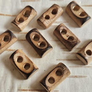 Puede incluir: Un conjunto de nueve botones de madera tipo palanca. Cada botón es rectangular con dos agujeros y un efecto de madera quemada marrón oscuro en los bordes. Los botones son de color marrón claro con veta natural.