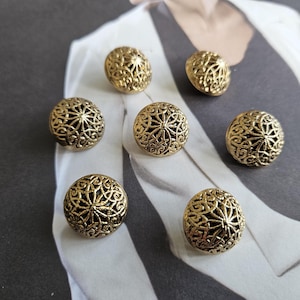 May include: A set of seven round, gold-toned buttons featuring an intricate, openwork filigree pattern. Each button has a domed, spherical shape with a detailed, repeating lattice design that creates a textured, metallic surface across the front.