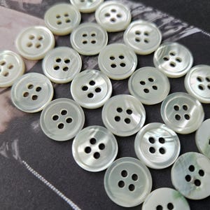 Può includere: Una collezione di bottoni rotondi bianchi perlati, ciascuno con quattro fori. I bottoni sono disposti su uno sfondo scuro, mettendo in risalto la loro sottile iridescenza e il design classico. Ideali per cucito e progetti artigianali.