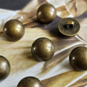 May include: Close-up of several antique brass-colored buttons. The buttons are round and have a slightly textured surface. One button has a loop on the back. The buttons are scattered on a patterned surface.