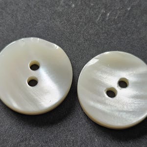 May include: Two round, off-white buttons with two holes each. The buttons have a pearlescent sheen and are set against a dark gray background. These buttons are suitable for sewing projects and garment construction.