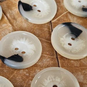 May include: A selection of round buttons, each with two holes. The buttons feature a cream-coloured base with a black and white marbled design. They are arranged on a brown, textured surface.