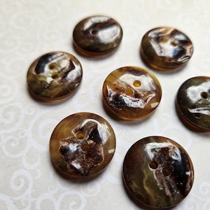 May include: Eight round buttons with two holes, in a marbled design. The buttons feature shades of brown, tan, and dark green. They are arranged on a light-coloured surface with a subtle pattern, suitable for sewing or craft projects.