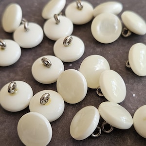 May include: A collection of round, off-white buttons with a smooth, glossy finish. Each button features a small, silver-toned metal shank on the back for attachment, arranged in a scattered pattern across the frame with a consistent circular shape.