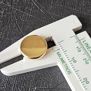 Könnte beinhalten: Ein kleiner, runder, goldfarbener Knopf mit einer glatten, reflektierenden Oberfläche. Der Knopf befindet sich zwischen den Backen einer weißen Kunststoff-Schieblehre, die einen Durchmesser von etwa 10 Millimetern anzeigt.