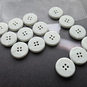 Peut inclure: Une collection de boutons ronds blanc cassé présentant un subtil motif marbré gris. Chaque bouton possède quatre trous centrés pour la fixation. Les boutons sont disposés de manière éparse, affichant une texture de surface lisse et mate.