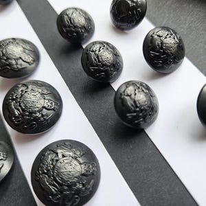 May include: A selection of dark grey, oval buttons featuring an intricate embossed design. The buttons are arranged on a black and white striped surface, providing a striking contrast. The buttons appear to be made of metal.