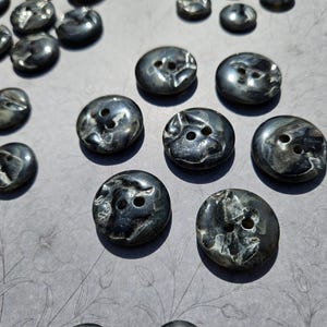 Peut inclure: Ensemble de boutons gris foncé à l'aspect marbré. Les boutons, de tailles variées, comportent deux trous de couture. Ils sont disposés sur une surface gris clair avec un motif floral subtil.