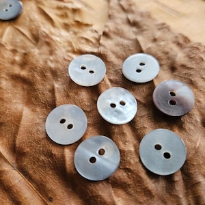 May include: A selection of round, pearlescent buttons, each with two holes, arranged on a textured, brown surface. The buttons have a subtle colour variation, with some appearing grey and others with a hint of brown. Suitable for sewing and craft projects.