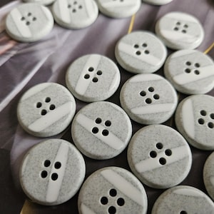 Puede incluir: Primer plano de varios botones redondos grises con cuatro agujeros y una franja diagonal blanca. Los botones tienen una textura moteada y están dispuestos en primer plano. Ideal para costura y manualidades.