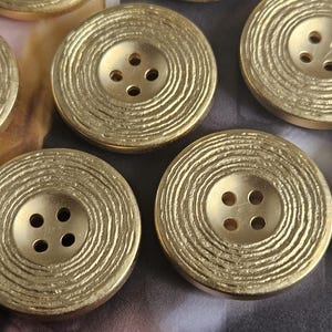 May include: Close-up of several gold-coloured buttons. Each button is round with a textured, concentric circle design and four holes for sewing. The buttons have a metallic sheen and a vintage aesthetic.