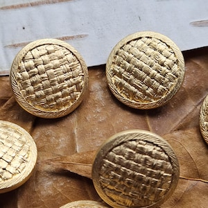 May include: Close-up of several gold-colored buttons with a woven texture. Each button is round with a textured border. The buttons are arranged on a brown leaf, creating a vintage or antique aesthetic. These buttons could be used for sewing or crafting projects.
