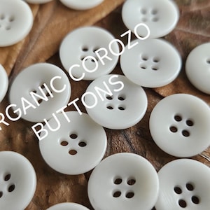May include: A collection of off-white, round corozo buttons with four holes each, scattered on a brown surface. The text "ORGANIC COROZO BUTTONS" is overlaid on the image in white. These buttons are ideal for sewing and craft projects.