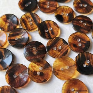 May include: A selection of round buttons featuring a tortoiseshell design. The buttons are a blend of amber and dark brown, each with two holes for sewing. The buttons are spread across a light-coloured surface.