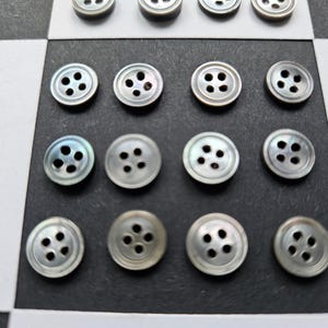 May include: A collection of iridescent, pearlescent buttons with four holes each, arranged on a black and white checkered surface. The buttons have a subtle sheen and are round in shape, suitable for sewing or craft projects.