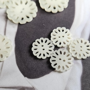 May include: Off-white, flower-shaped buttons with scalloped edges and two sewing holes. The buttons are scattered on a textured surface. The buttons are approximately 2.5 cm in diameter.
