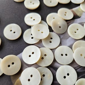 Puede incluir: Un conjunto de botones redondos de color blanco roto, cada uno con dos agujeros. Los botones están dispersos sobre una superficie estampada, mostrando su textura suave y ligeramente iridiscente. Ideal para costura y manualidades.