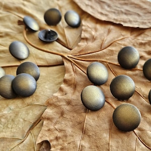 May include: A selection of antique brass-coloured buttons with a domed shape, scattered on a dried, brown leaf. The buttons have a metallic sheen and a slightly aged appearance, suggesting vintage or rustic design elements.