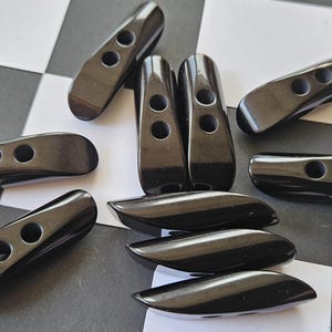 May include: A collection of ten elongated, glossy black toggle buttons, each featuring two circular holes for attachment. These smooth, curved fasteners are arranged in a scattered pattern, showcasing their uniform dark finish and distinct shape.
