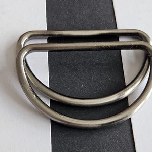 May include: Two metallic D-rings with a smooth, gunmetal gray finish. Each ring features a flat straight edge and a curved semi-circular bottom, designed for use as a hardware component in various sewing, bag making, or strap assembly projects.
