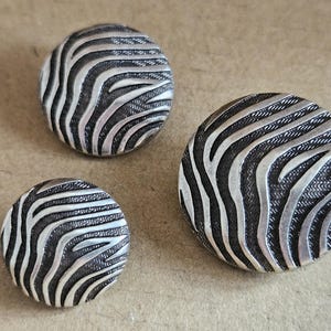 May include: Three circular buttons of varying sizes featuring a raised silver-toned zebra stripe pattern against a recessed, dark, textured background. The metallic finish highlights the curved surface of each button, creating a distinct contrast.