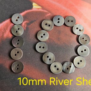 Op de afbeelding: Een verzameling van 10 mm rivierschelpknoppen. De knopen zijn rond, grijs en hebben twee gaten om op te naaien. De knopen zijn gerangschikt in een cirkel en een verticale lijn. De tekst "10mm River Shell" wordt in het geel weergegeven.