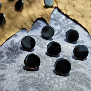 May include: A collection of small, circular, black buttons with a smooth, glossy finish. The buttons are arranged in a cluster, featuring a simple, flat, and round design. Each button has a reflective surface that catches the light, highlighting the uniform dark colour and the clean, circular edges of the individual pieces.