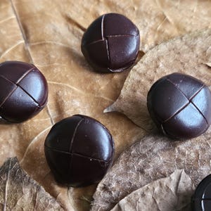 May include: Five dark brown leather buttons with a cross-stitch pattern are displayed. The buttons are spherical and have a textured surface. They are placed on a bed of dried brown leaves.