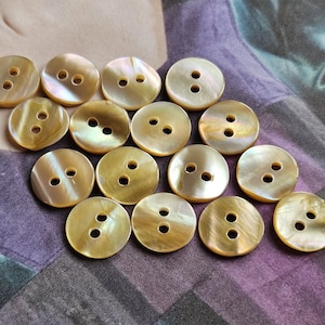 May include: A collection of round, iridescent buttons with two holes each. The buttons have a pearlescent sheen and a warm, golden hue. They are arranged on a patterned fabric background, suggesting a craft or sewing project.