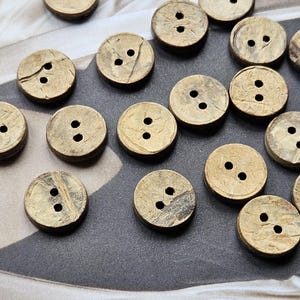 Puede incluir: Un conjunto de botones redondos de cáscara de coco de color natural, con dos o tres agujeros. Los botones tienen una superficie texturizada y están dispuestos sobre un fondo estampado, ideales para coser y manualidades.