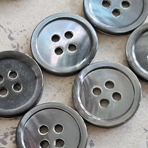May include: A collection of round, dark gray buttons, each featuring a smooth, slightly iridescent surface and four centered holes for attachment. The buttons have a raised outer rim and a polished finish, arranged in a scattered pattern.