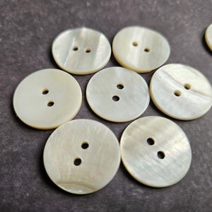Op de afbeelding: Een verzameling ronde, crèmekleurige knopen met elk twee gaatjes. De knopen hebben een parelmoerglans en liggen op een donkere, getextureerde ondergrond. Ideaal voor naai- en knutselprojecten.