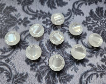 May include: Nine round, iridescent mother-of-pearl buttons with a textured outer ring and a smooth center. The buttons are arranged on a gray and white floral patterned background.