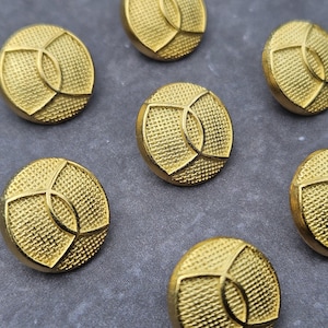 Könnte beinhalten: Nahaufnahme von mehreren goldfarbenen Knöpfen. Jeder Knopf ist rund mit einer strukturierten Oberfläche und einem geometrischen Design in der Mitte. Die Knöpfe sind auf einer grauen Oberfläche verstreut.