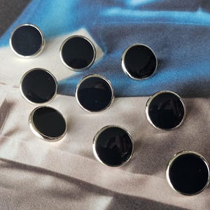 May include: A collection of ten round buttons with a glossy black center and a silver-colored metal rim. The buttons are scattered on a patterned fabric surface, creating a visually appealing arrangement. These buttons are suitable for various sewing projects.