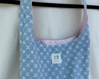 以下が含まれることがあります： 白の花柄が施された水色のトートバッグ。バッグの内側はピンク色で、長いストラップが付いています。バッグは黒いハンガーに掛けられています。バッグの前面には小さなラベルが縫い付けられています。