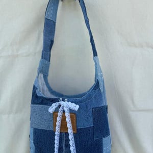Könnte beinhalten: Schultertasche aus Patchwork-Denim in verschiedenen Blautönen. Die Tasche hat ein geschwungenes Design, eine weiße Spitzenschleife und ein braunes Lederetikett. Sie hat einen langen Riemen und hängt vor einem weißen Hintergrund. Ein stilvolles Accessoire.