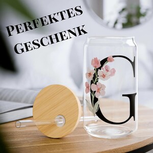 Könnte beinhalten: Ein klares, dosenförmiges Trinkglas mit Bambusdeckel und Strohhalm. Das Glas hat einen schwarzen Buchstaben "G" mit einem floralen Design in Rosa und Weiß. Der Text "PERFEKTES GESCHENK" steht über dem Glas.
