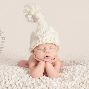 May include: A newborn baby wearing a white knitted hat with a pom-pom on top, sleeping on a white fluffy blanket.