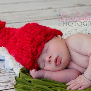 May include: A newborn baby wearing a red knitted hat with a white pom-pom sleeps on a green fabric. The baby is lying on its side with its arm under its head. The hat is covering the baby's eyes.