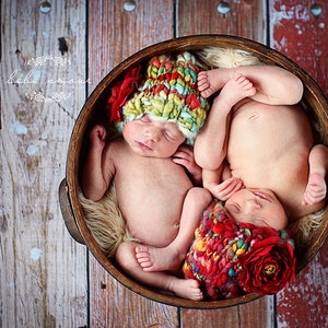 May include: Two newborn babies sleeping in a wooden bowl, wearing colourful crocheted hats with red flower accents. The babies are nestled in a soft, white fur lining.