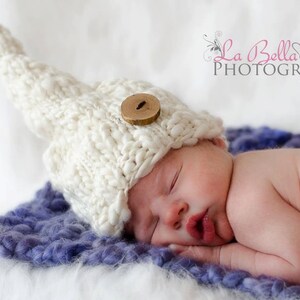 May include: A newborn baby wearing a white knitted hat with a wooden button, sleeping on a purple fluffy blanket. The baby is curled up on its side with its hand under its chin.