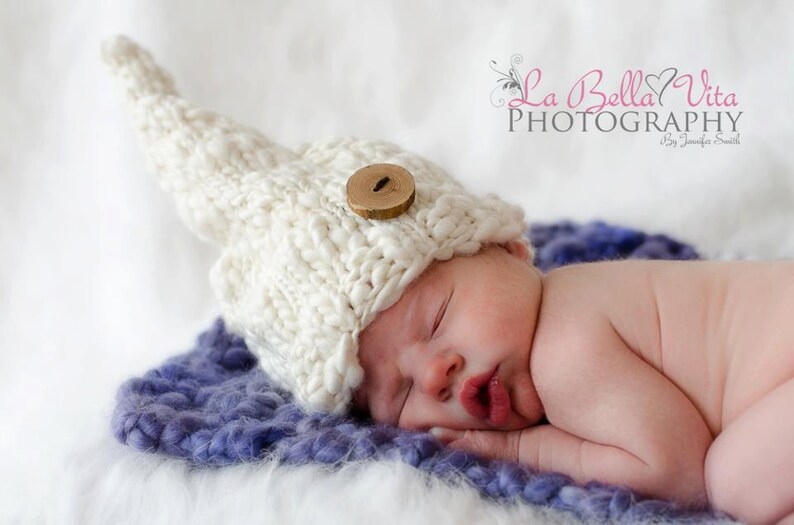 May include: A newborn baby wearing a white knitted hat with a wooden button, sleeping on a purple fluffy blanket. The baby is curled up on its side with its hand under its chin.