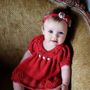 Op de afbeelding: Een babymeisje in een rode gebreide jurk met een witte kraag en blauwe knopen. Ze draagt een rood en wit hoofdband met bloemen. De baby zit in een bruine fauteuil.