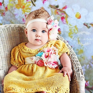 Op de afbeelding: Een babymeisje in een gele gebreide jurk met een bloemenversiering en een hoofdband met een bloemenaccent. De baby zit in een witte rieten stoel tegen een bloemige achtergrond.