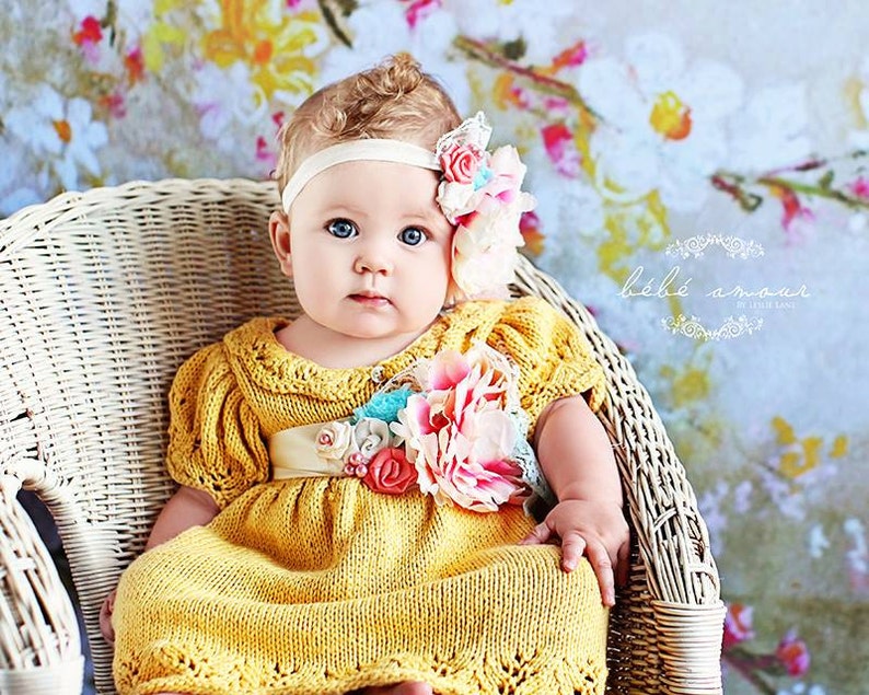 Op de afbeelding: Een babymeisje in een gele gebreide jurk met een bloemenversiering en een hoofdband met een bloemenaccent. De baby zit in een witte rieten stoel tegen een bloemige achtergrond.