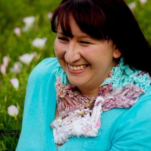 May include: A woman with brown hair is smiling and wearing a turquoise blue shirt with a white and pink crocheted scarf. The scarf is made of different textures and colors.