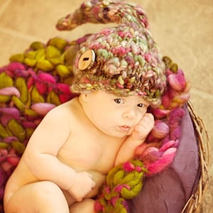 May include: A baby wearing a multi-coloured crocheted hat with a wooden button. The baby is lying in a basket filled with colourful yarn.