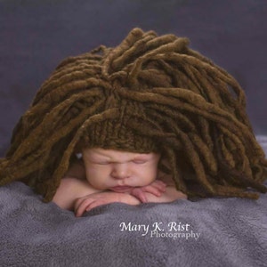 May include: A newborn baby wearing a brown knitted hat with long brown yarn dreadlocks. The baby is lying on a grey blanket and has their eyes closed.