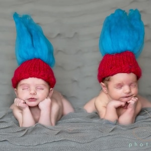 May include: Two newborn babies wearing red knit hats with blue yarn hair, sleeping on a gray textured blanket.